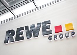 Logo der REWE Group mit grauer Schrift und roten sowie gelben Quadraten an einer Wand.